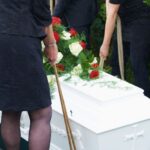 Pessoas segurando cordas descem caixão branco para expor custos invisíveis do sepultamento tradicional.